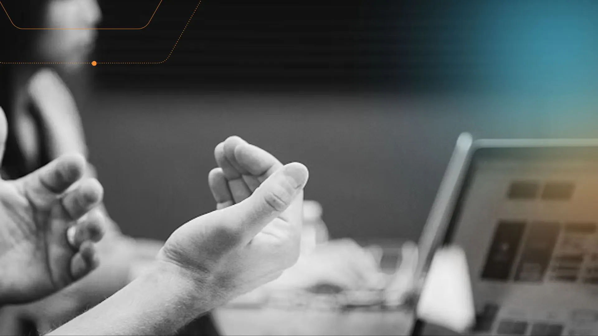 two people sat at desk, blurred, with hands in foreground and laptop in background